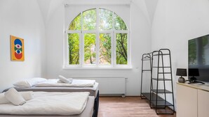 A modern hotel room with two beds, a large arched window, a radiator, a black metal shelf unit, a television, and a framed artwork on the wall. - EXQUIS Luxus 3-Room Maisonette I Stuttgart West Schwabstraße Train Station (Stuttgart)