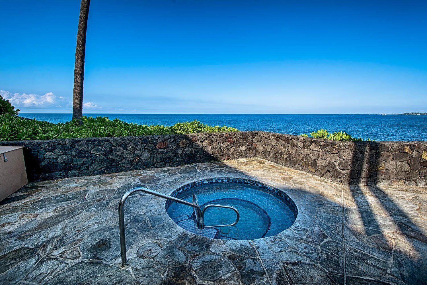 A stone pool with a metal ladder, surrounded by a stone wall and greenery, overlooking a clear blue ocean.