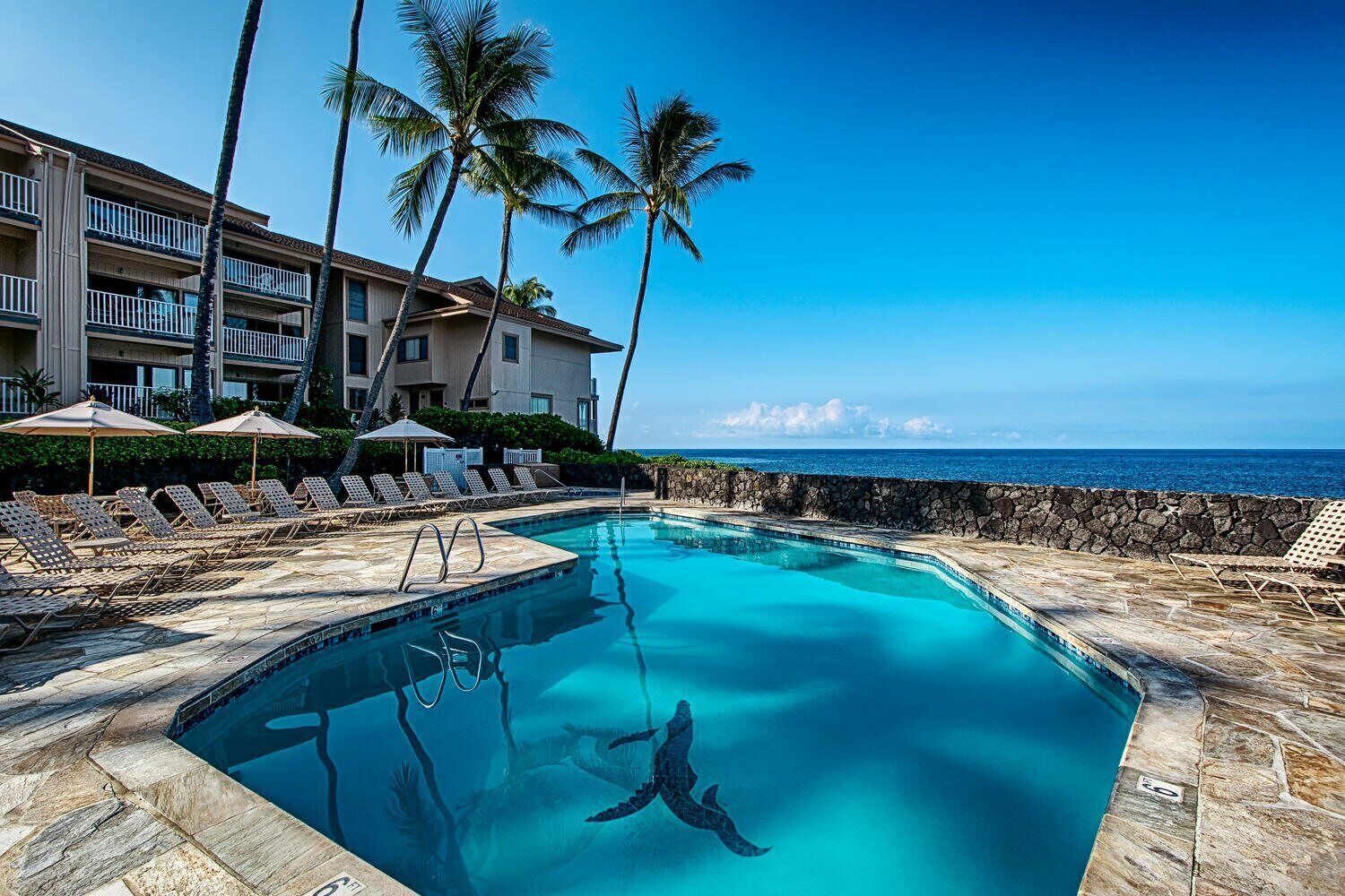 A pool with clear blue water, surrounded by lounge chairs and umbrellas, with a view of the ocean and a building in the background.