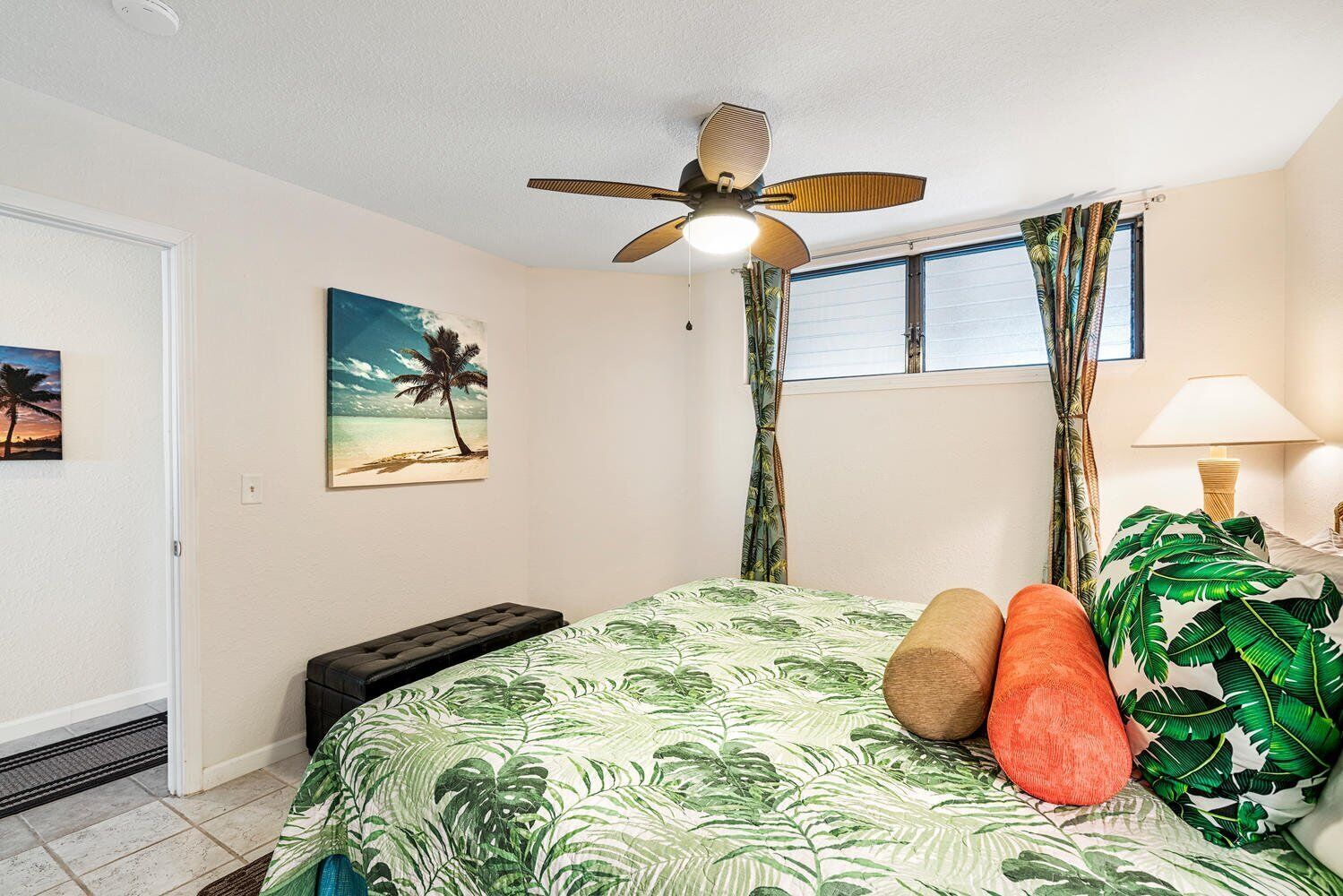 A bedroom with a bed, a ceiling fan, a window with curtains, a nightstand with a lamp, and a door.