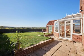 A house with a brick exterior, a glass door leading to a patio, and a view of a green lawn and distant hills. - Host & Stay | Cherry Willows (Stainsacre)