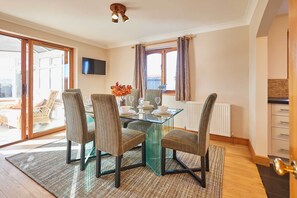 A dining area with a glass table, four chairs, a vase of flowers, and a television on the wall. - Host & Stay | Cherry Willows (Stainsacre)