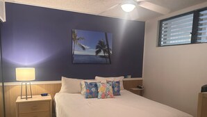 A bedroom with a bed, a nightstand, a lamp, a painting of a beach scene, and a window with blinds. - Top Floor Nice Ocean View Close to Town! - Vrkcv (Kailua-Kona)