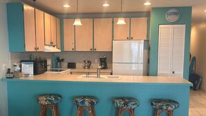 A kitchen with teal cabinets, a white refrigerator, a sink, and three bar stools with floral cushions. - Top Floor Nice Ocean View Close to Town! - Vrkcv (Kailua-Kona)