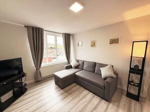 Ein modernes Wohnzimmer mit einem Ecksofa, einem Flachbildfernseher, einem Fenster mit Vorhängen und einem kleinen Regal.