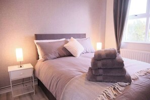 Ein ordentlich bezogenes Bett mit Kopfteil, einer Nachttischlampe, einem Stapel Handtücher und einem Fenster mit Vorhängen.