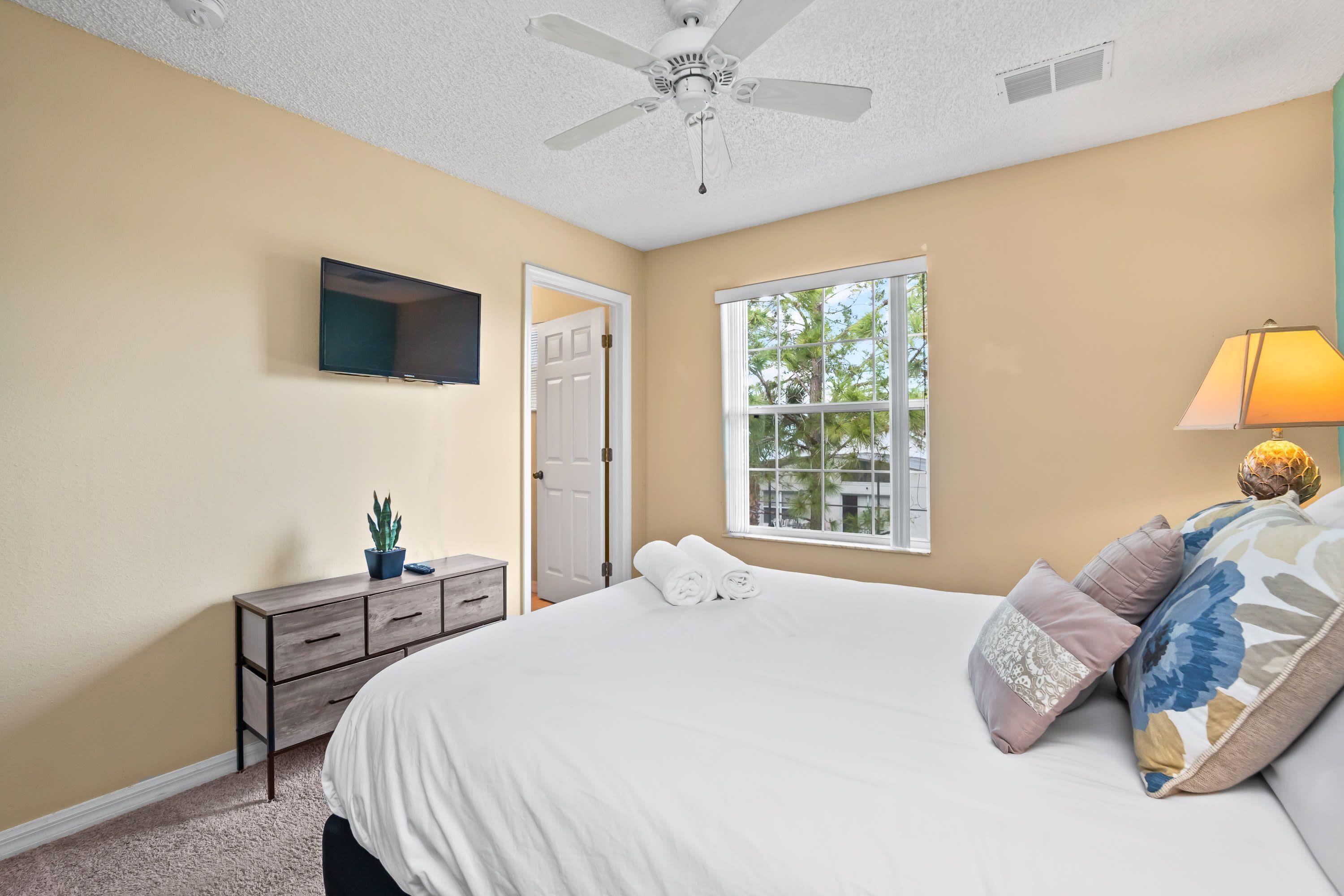 A bedroom with a bed, a ceiling fan, a TV, a window, and a lamp.