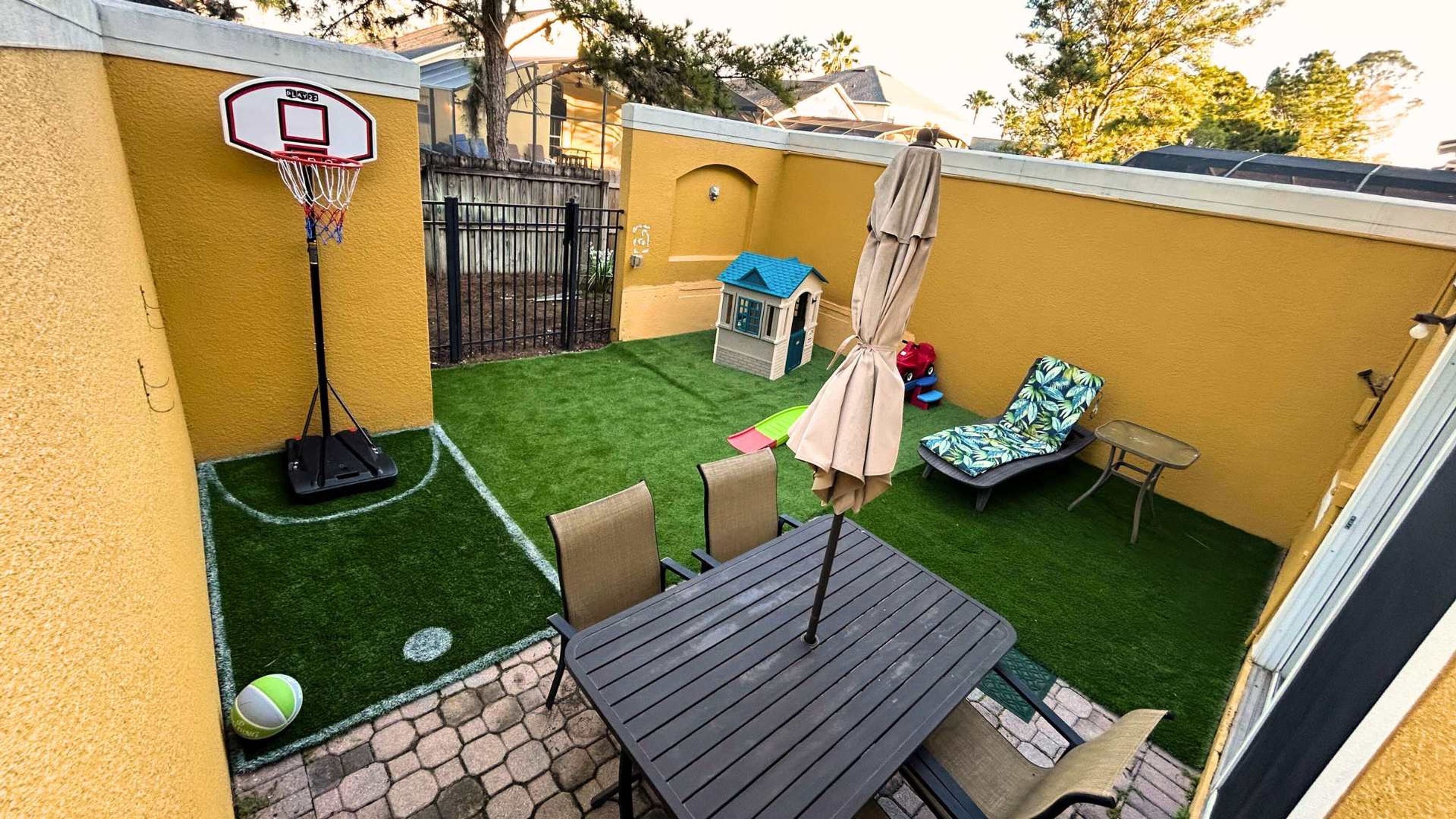 A backyard with artificial turf, a basketball hoop, a small playhouse, and a lounge chair.