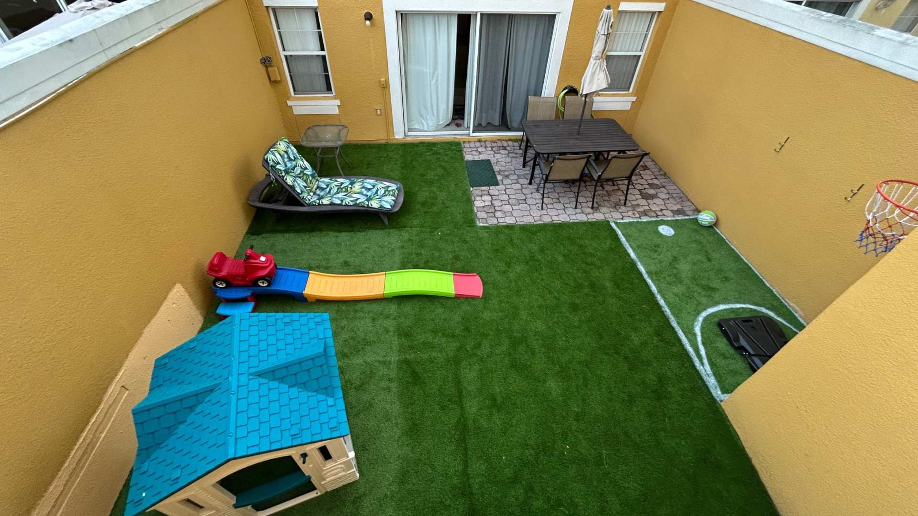A small backyard with artificial grass, a play structure, a table with chairs, and a basketball hoop.