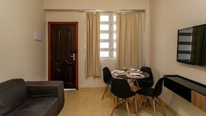 A small, furnished room with a dining table, four chairs, a sofa, a TV, and a window with curtains. - Copacabana - O coraçao do Rio (Rio de Janeiro)