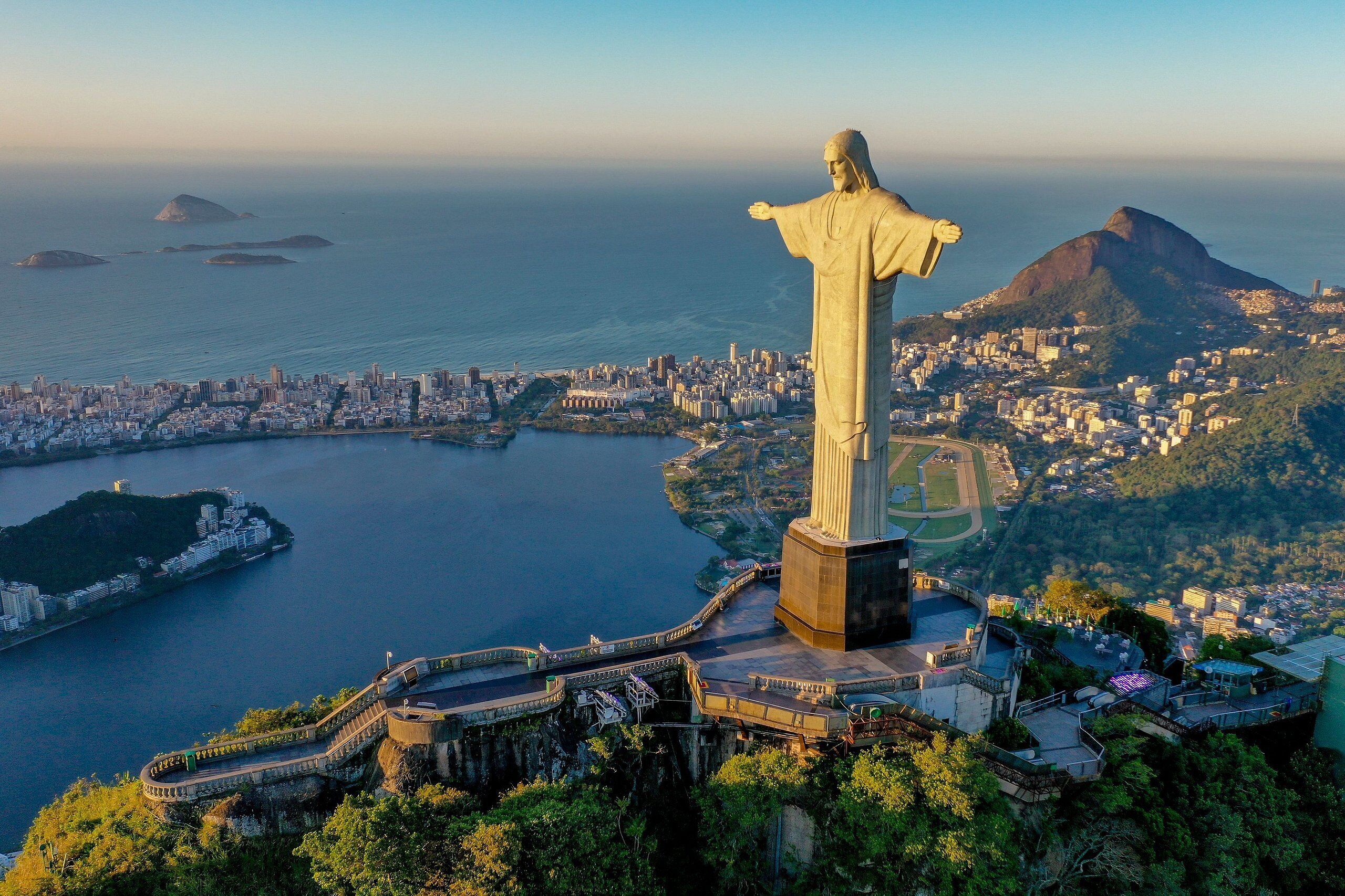 A large statue overlooking a coastal city with a bridge and lush greenery.