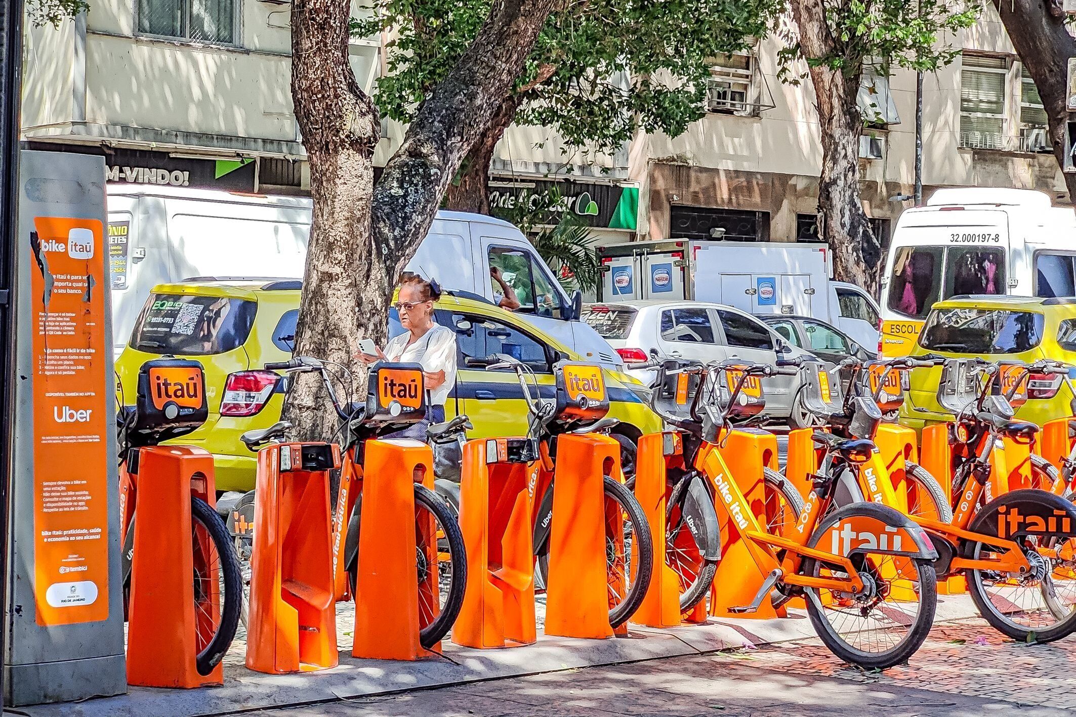Eine Reihe von orangefarbenen Fahrradstellern mit 'itau'-Branding, ein gelbes Taxi und ein weißer Transporter in der Nähe.