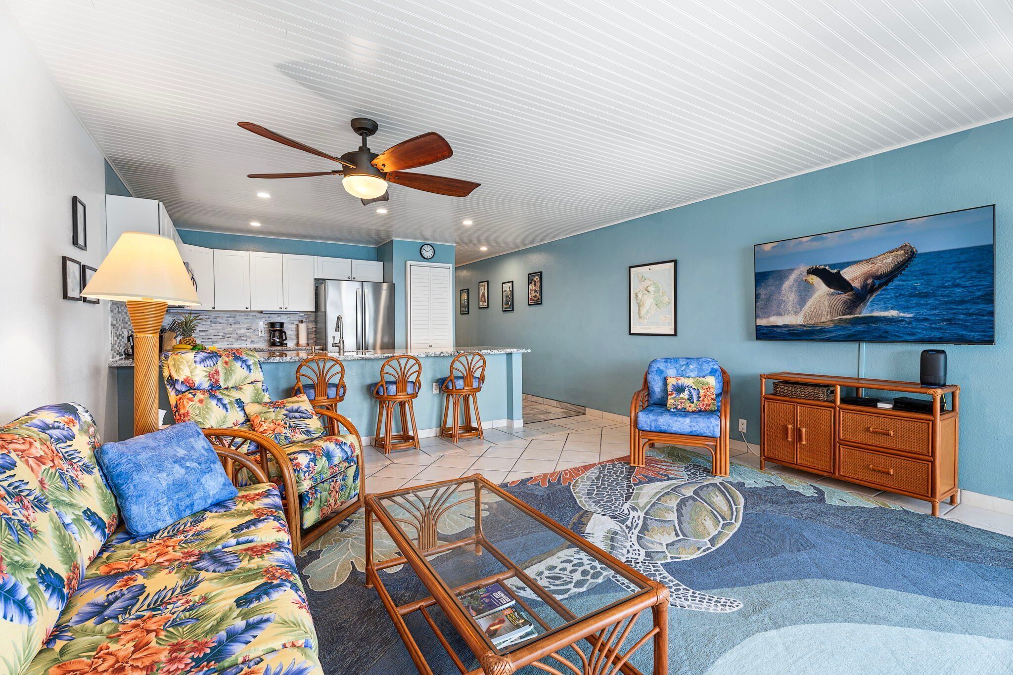 A living room with a sofa, chairs, a coffee table, a ceiling fan, a kitchen area, and a large whale mural on the wall.