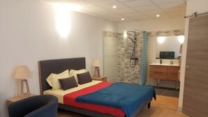 A bedroom with a bed, bedside tables, a chair, a shower area, and a bathroom with a sink and mirror.