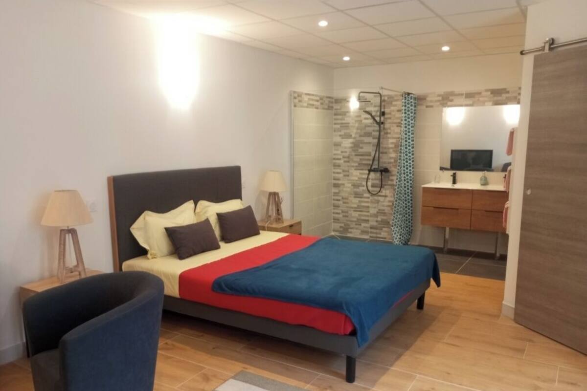 Een slaapkamer met een bed, nachtkastjes, een stoel, een douche en een badkamer met een wastafel en spiegel.