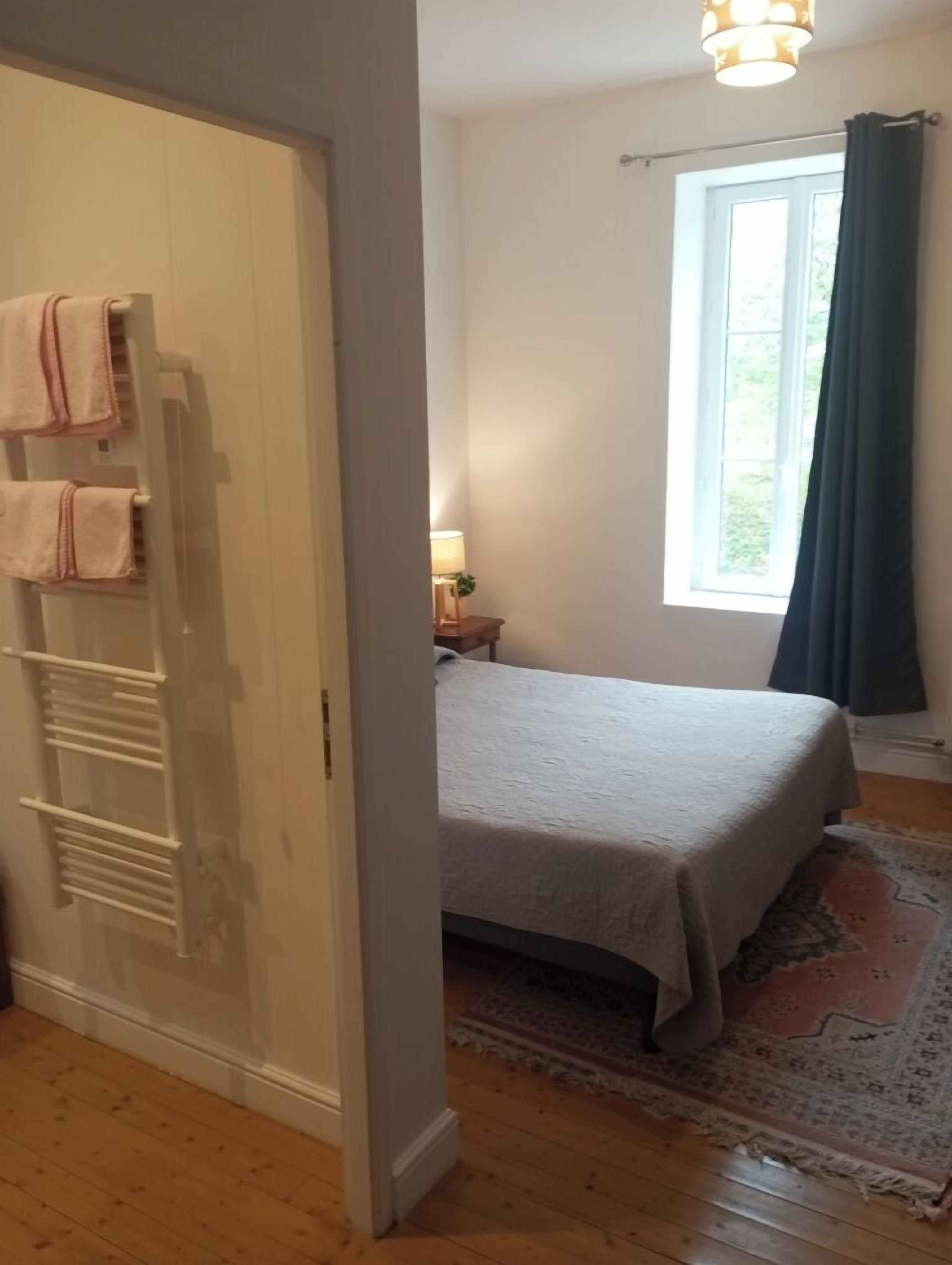 Ein Schlafzimmer mit Bett, einem Fenster mit Vorhängen, einem Heizkörper und einer Tür mit daran hängenden Handtüchern.