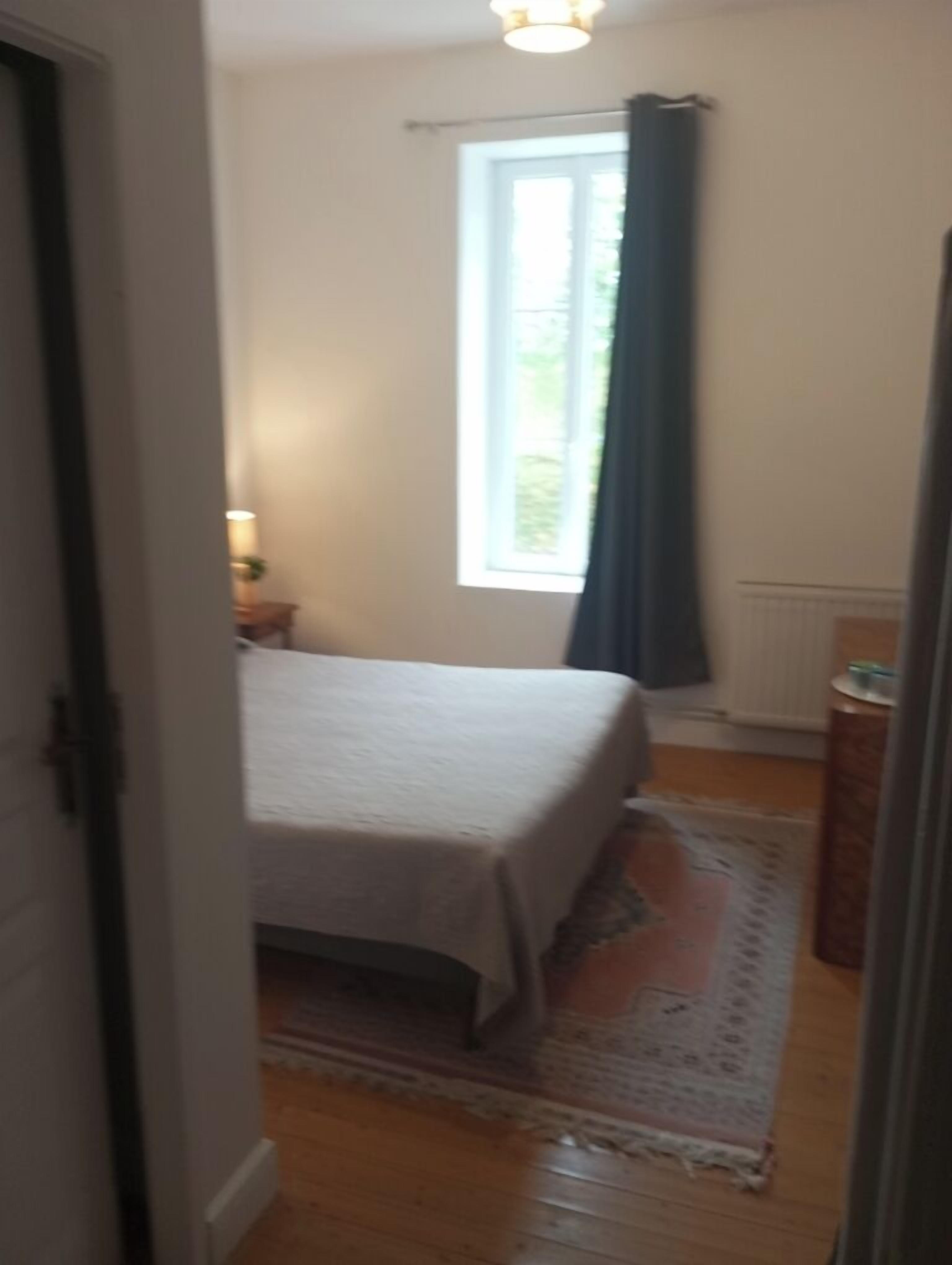 A bedroom with a bed, a window with curtains, a radiator, and a small table with a lamp.