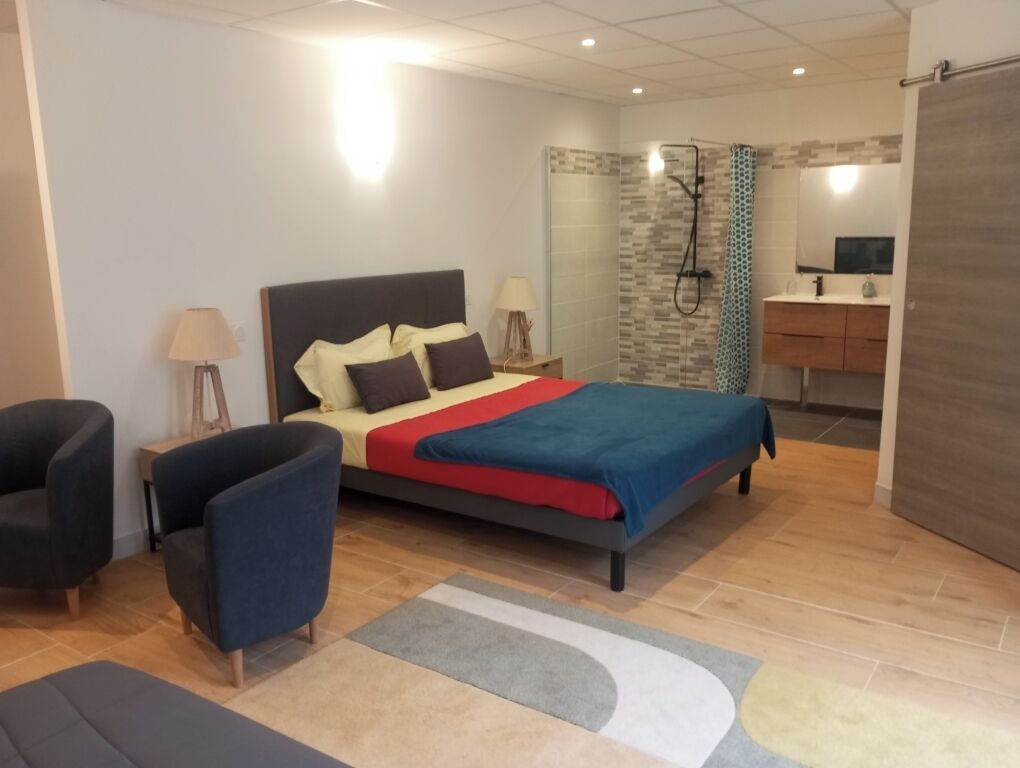 A modern bedroom with a bed, bedside tables, a chair, a bathroom with a shower, and a sink.