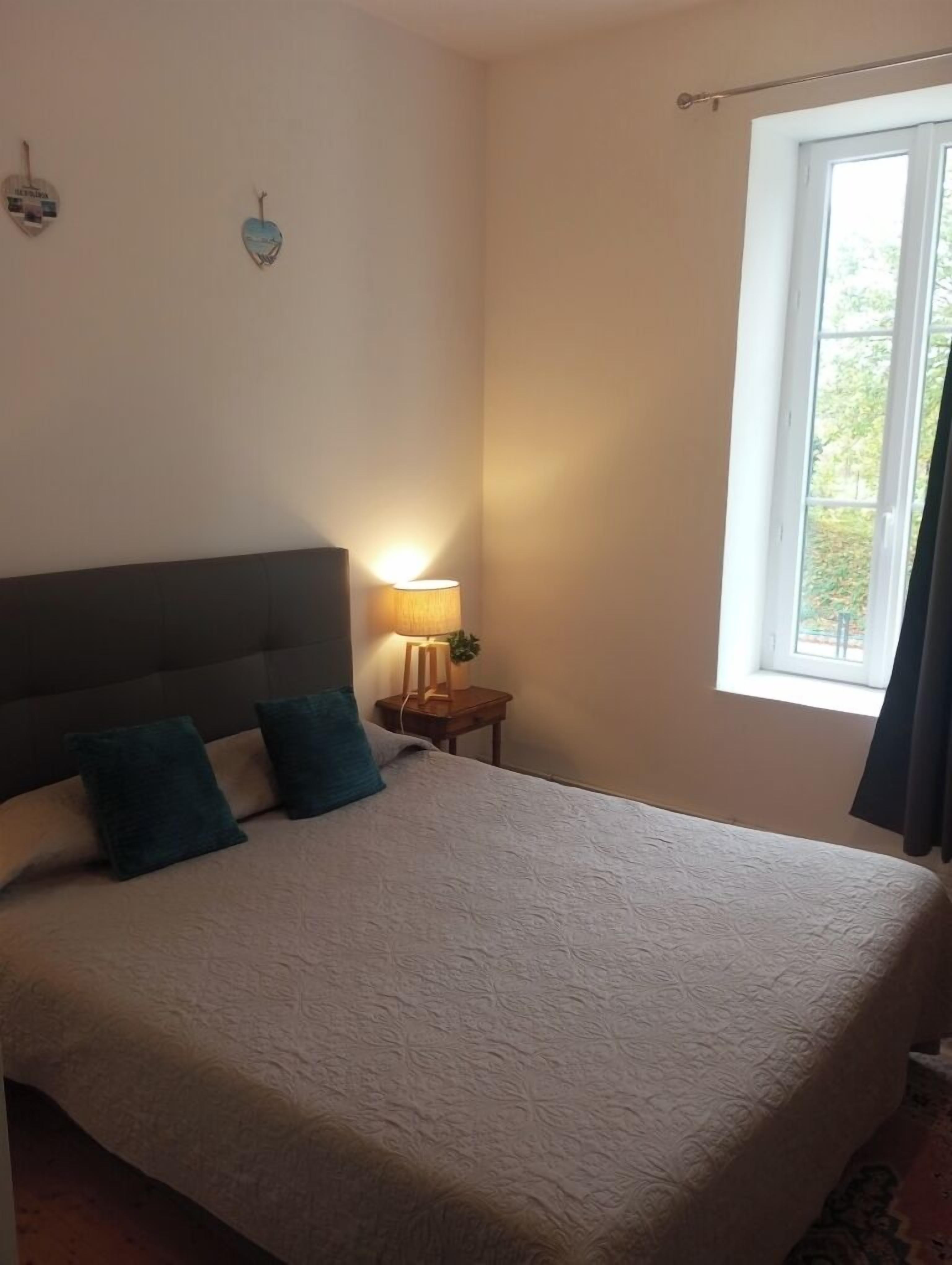Cama bem arrumada com cabeceira, luminária de cabeceira e janela com cortinas.