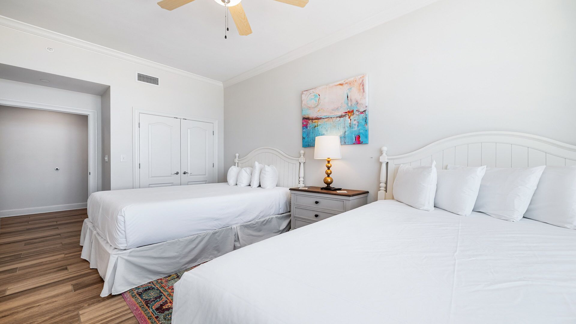 A hotel room with two single beds, a nightstand with a lamp, a ceiling fan, and a painting on the wall.