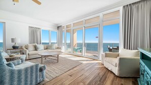 A spacious living room with a large window overlooking the ocean, a sofa, a coffee table, and a view of the balcony. - Penthouse 4BR, Beachfront!, Lazy River/Slide! (Orange Beach)