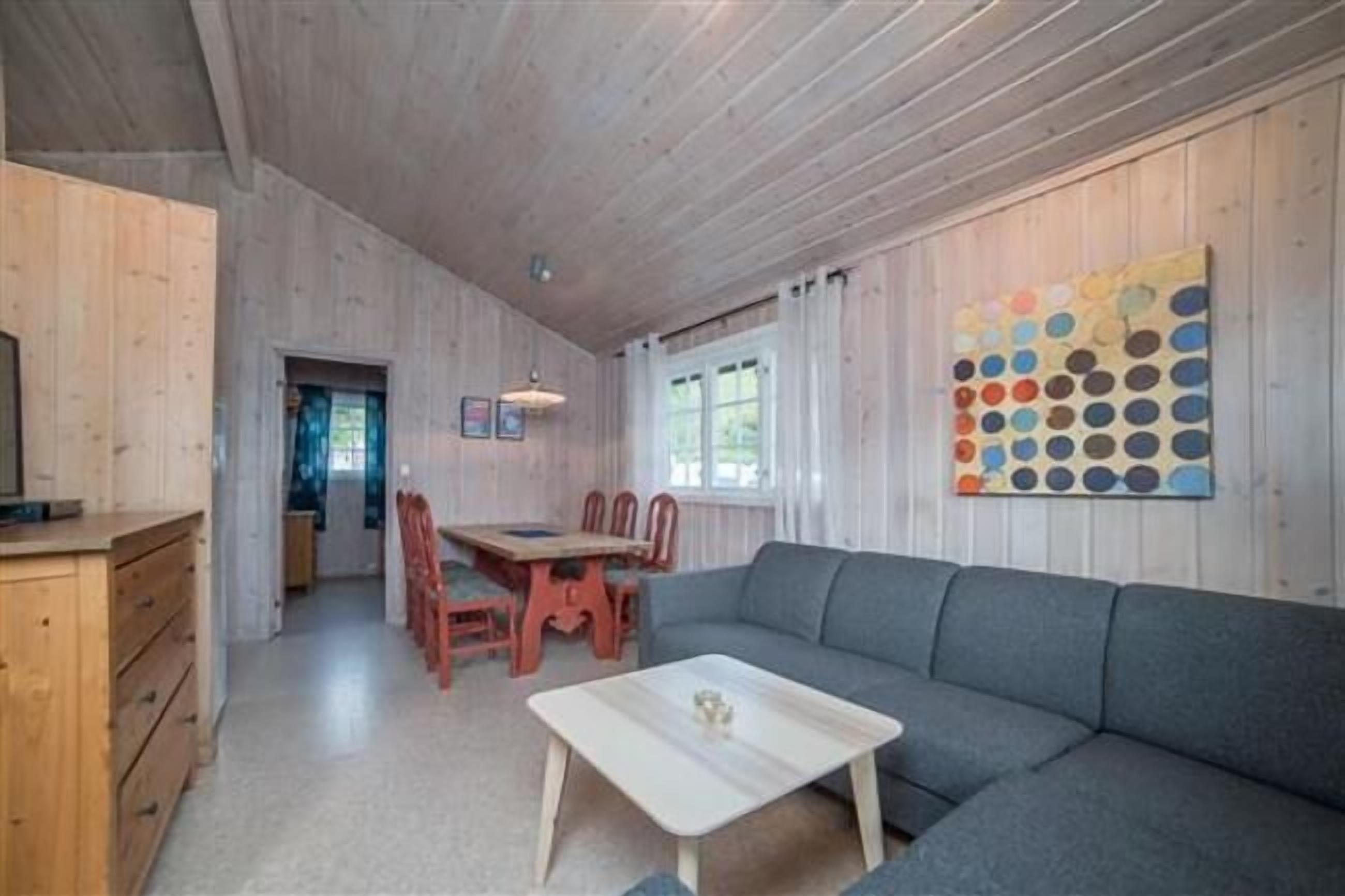 A cozy room with a wooden ceiling, a gray sofa, a wooden dining table with chairs, and a colorful wall art piece.