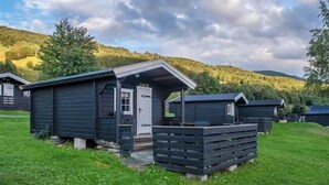A row of small, dark-colored cabins with a simple design, set in a grassy area with a backdrop of hills and a partly cloudy sky. - Pluscamp Mageli  (Øyer)