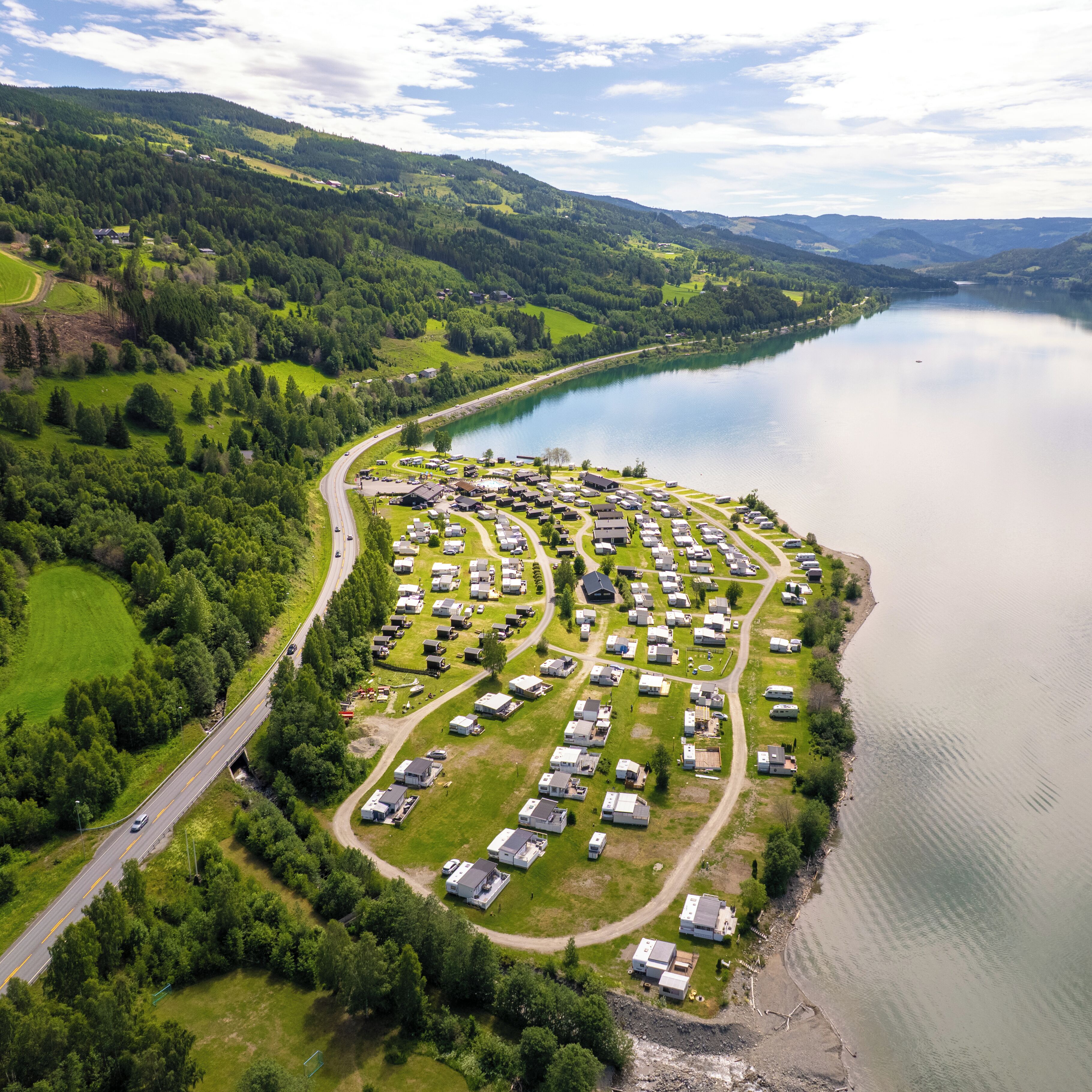 An aerial view of a lakeside campground with RVs and cabins, surrounded by lush greenery and hills.