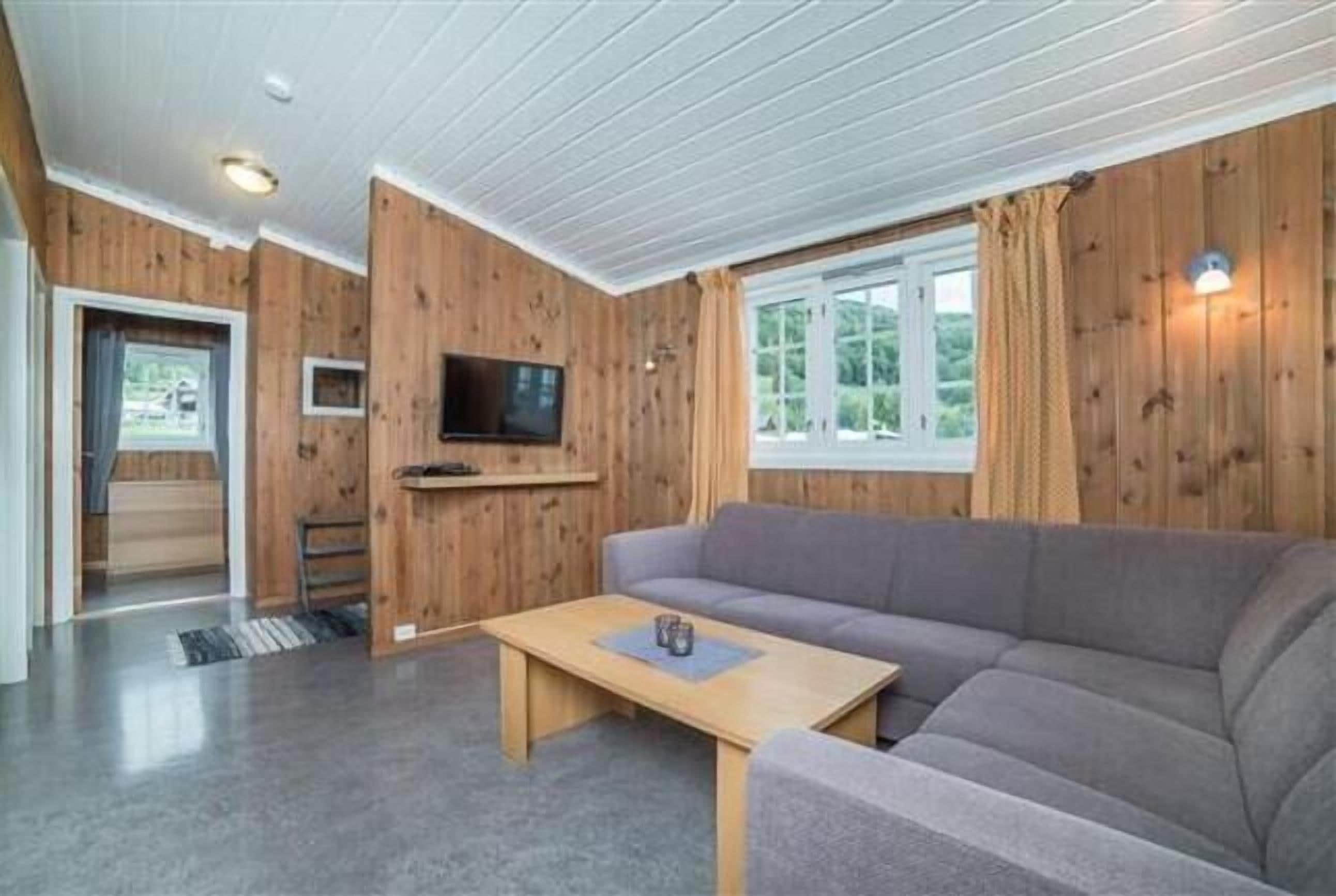 Une pièce chaleureuse avec un décor intérieur en bois, un canapé gris, une table basse en bois, un téléviseur à écran plat fixé au mur et une fenêtre avec des rideaux.