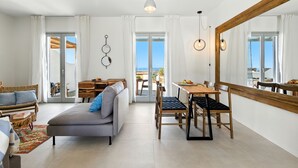 A modern living room with a sofa, dining table, and chairs. There is a large mirror on the wall and a view of the outdoors through the windows. - Selinofos - a modern seaside home blending comfort and sea views. (Naxos)