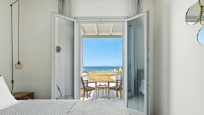 A bedroom with a large bed, a nightstand, a table with chairs, and a view of the beach through an open door. - Selinofos - a modern seaside home blending comfort and sea views. (Naxos)