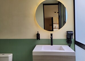 Ein Badezimmer mit einem weißen Waschbecken, einem runden Spiegel mit Beleuchtung, einem schwarzen Wasserhahn, einer Seifenspender und einer Toilette.