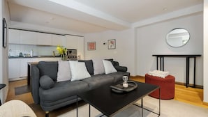 A modern living room with a grey sofa, a black coffee table, and a red ottoman. A kitchen is visible in the background with white cabinets and a tiled backsplash. - w Awesome 2BR in Roma Norte (Mexico City)
