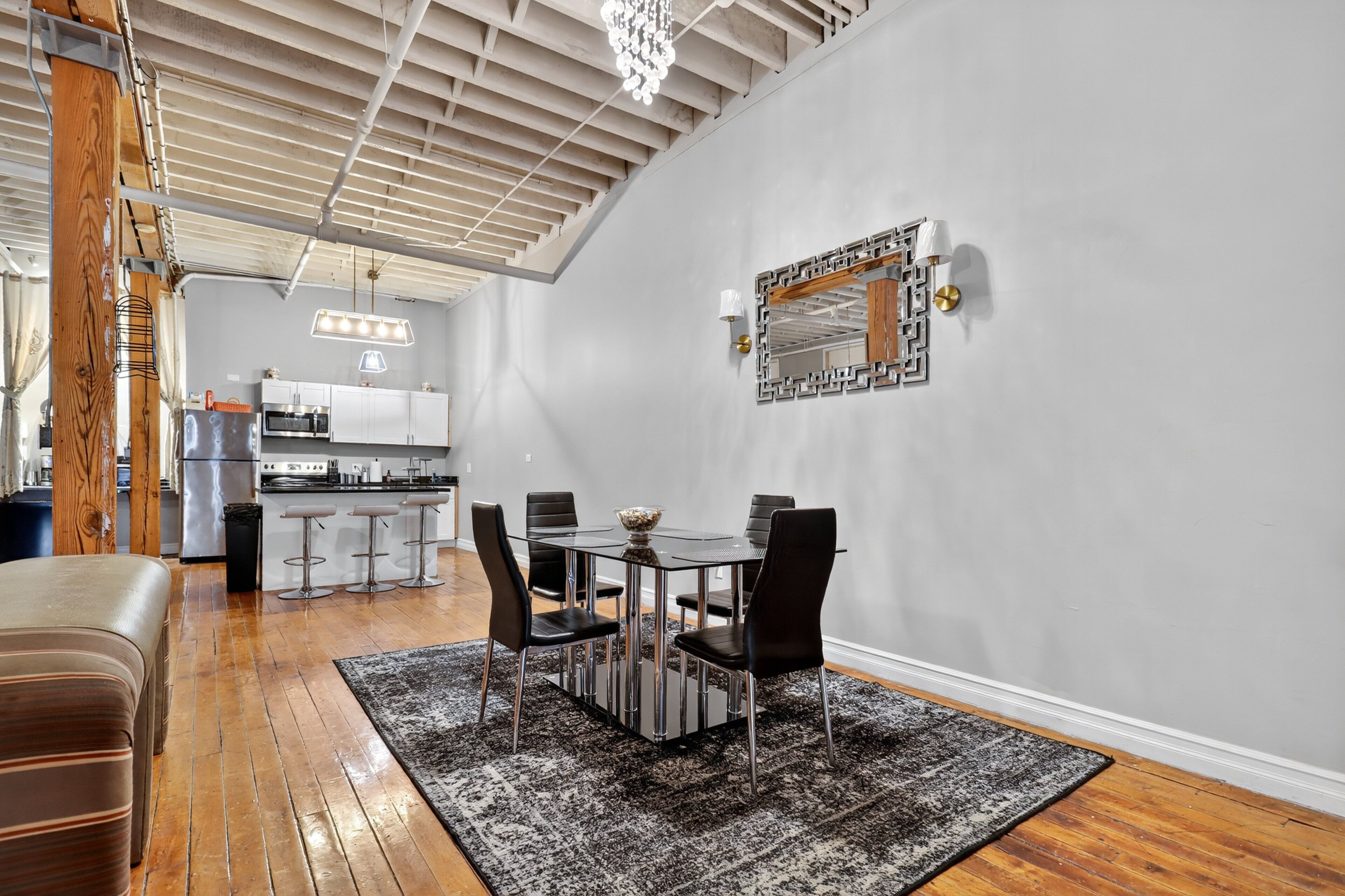 Una cocina moderna con electrodomésticos de acero inoxidable, un comedor con mesa de vidrio y sillas negras, y una pared gris con un espejo decorativo.