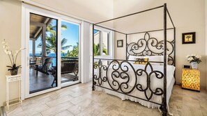 A four-poster bed with a balcony view, a small bedside table, and a nightstand with a clock. - Oceanfront Corner Penthouse with Sunset Views, Partial A/C, and Wraparound Lanai (Kailua-Kona)