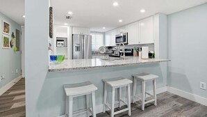A modern kitchen with white cabinets, a stainless steel refrigerator, a microwave, and a granite countertop. There are three white bar stools and a window with blinds. - Oceanfront Condo, Pool Access & Private Patio (St. Augustine)