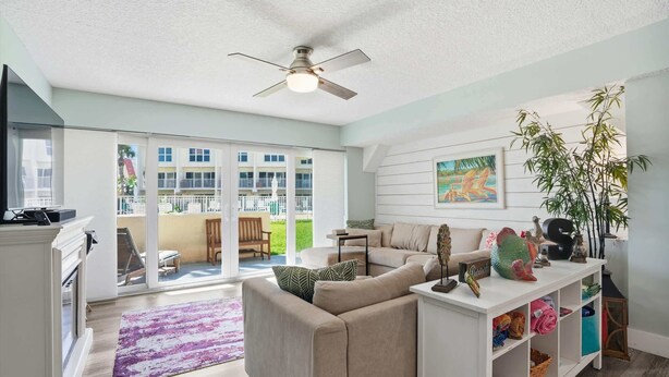 A spacious living room with a large window, a ceiling fan, a sofa, a coffee table, and a bookshelf. - Oceanfront Condo, Pool Access & Private Patio (St. Augustine)