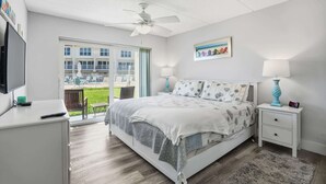 A bedroom with a bed, bedside tables, a ceiling fan, a TV, and a window with a view of a building and a lawn. - Oceanfront Condo, Pool Access & Private Patio (St. Augustine)