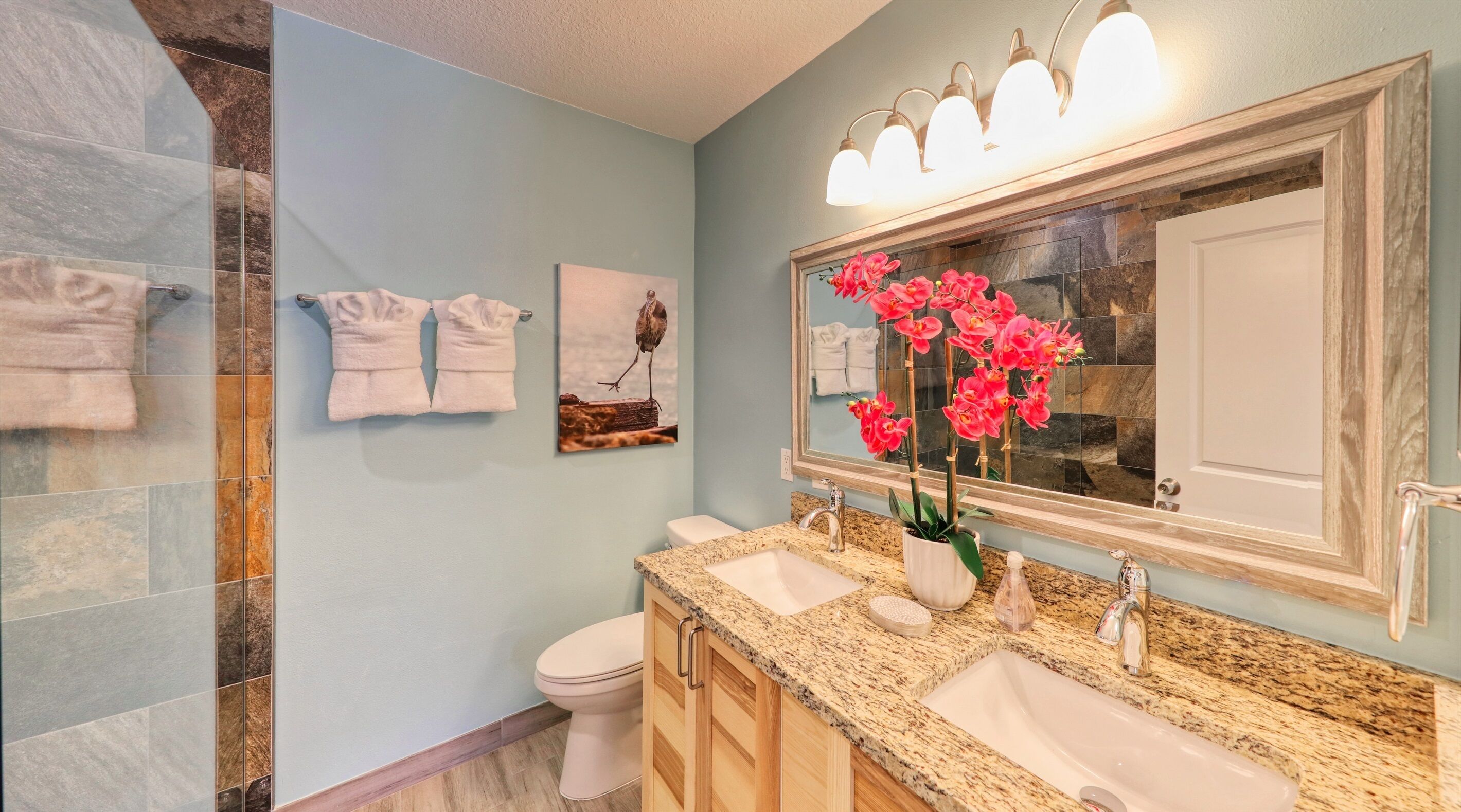 A bathroom with a light blue wall, a large mirror, two sinks, a toilet, and a shower area.