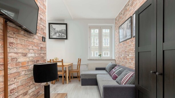 Ein gemütliches Wohnzimmer mit Backsteinwand, Flachbildfernseher, grauem Sofa, Holztisch und Fenster mit weißen Rahmen.