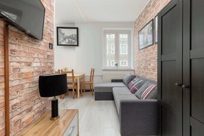 A cozy living room with a brick wall, a flat-screen TV, a grey sofa, a wooden table, and a window with white frames. - Spacious Apartment for 8 Persons | Heart of the Old Town (Gdańsk)