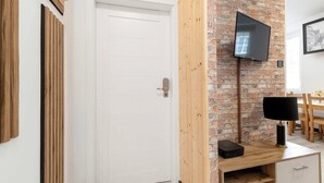 Ein modernes Zimmer mit weißer Tür, einer Ziegelwand, einem an der Wand befestigten Fernseher, einem Holztischchen mit Lampe und einem kleinen Schrank.