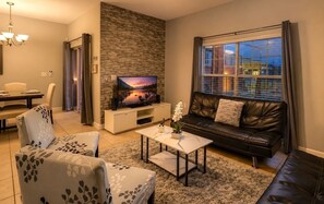 A modern living room with a flat-screen TV, a leather sofa, a dining table with chairs, and a large window with curtains. - Renovated 4BR Paradise Palms Resort Pool Townhome near Disney (Kissimmee)
