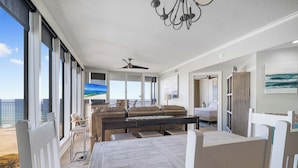 A spacious living area with a large window overlooking the beach, a ceiling fan, and a dining table with chairs. - The Colonnades 701 by Liquid Life (Gulf Shores)