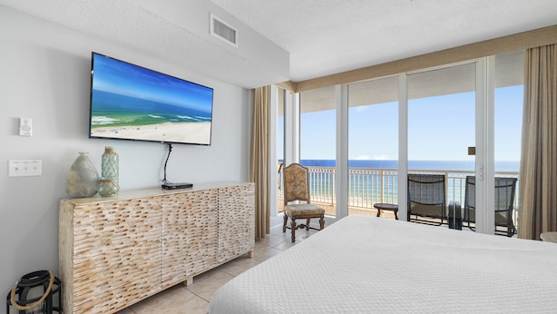 A hotel room with a large TV, a wooden cabinet, a bed, and a balcony with a view of the beach. - The Colonnades 701 by Liquid Life (Gulf Shores)