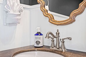 Un baño con un lavamanos blanco, un espejo con marco dorado y una botella de jabón Beach House.