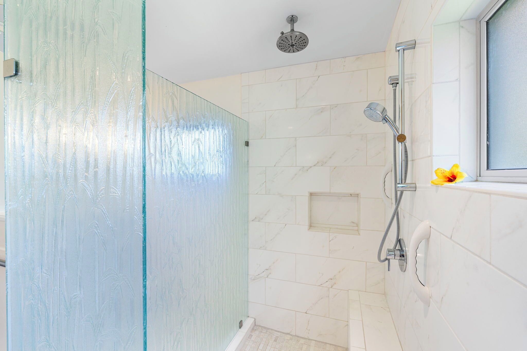 A bathroom with a glass shower enclosure, a handheld showerhead, a wall-mounted faucet, and a window with a flower decoration.