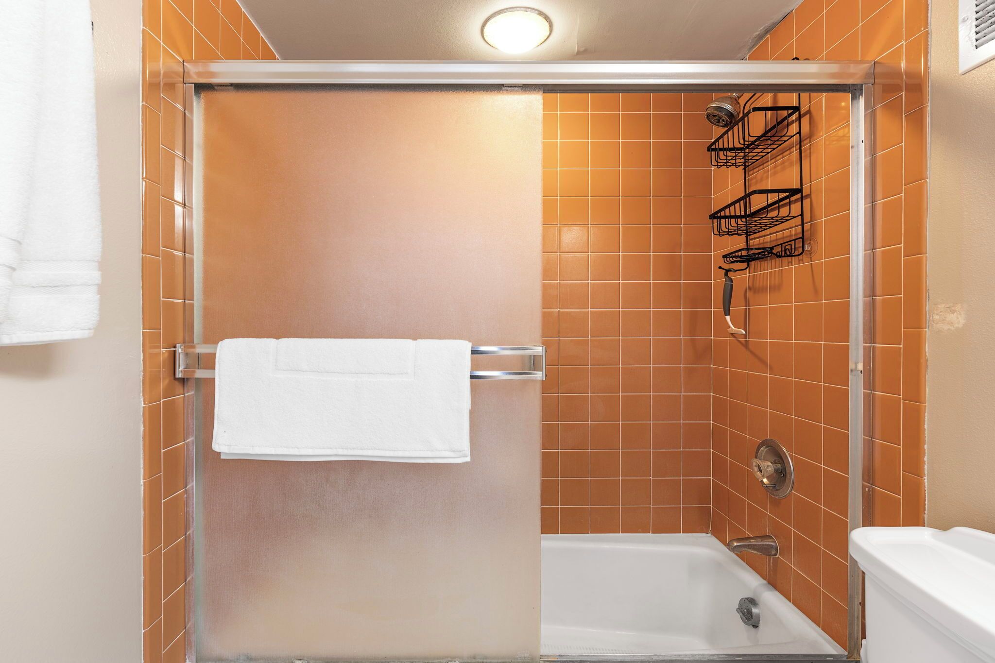 Un área de ducha con puerta de cristal, una toalla blanca colgada en un perchero metálico y un cabezal de ducha montado en la pared.