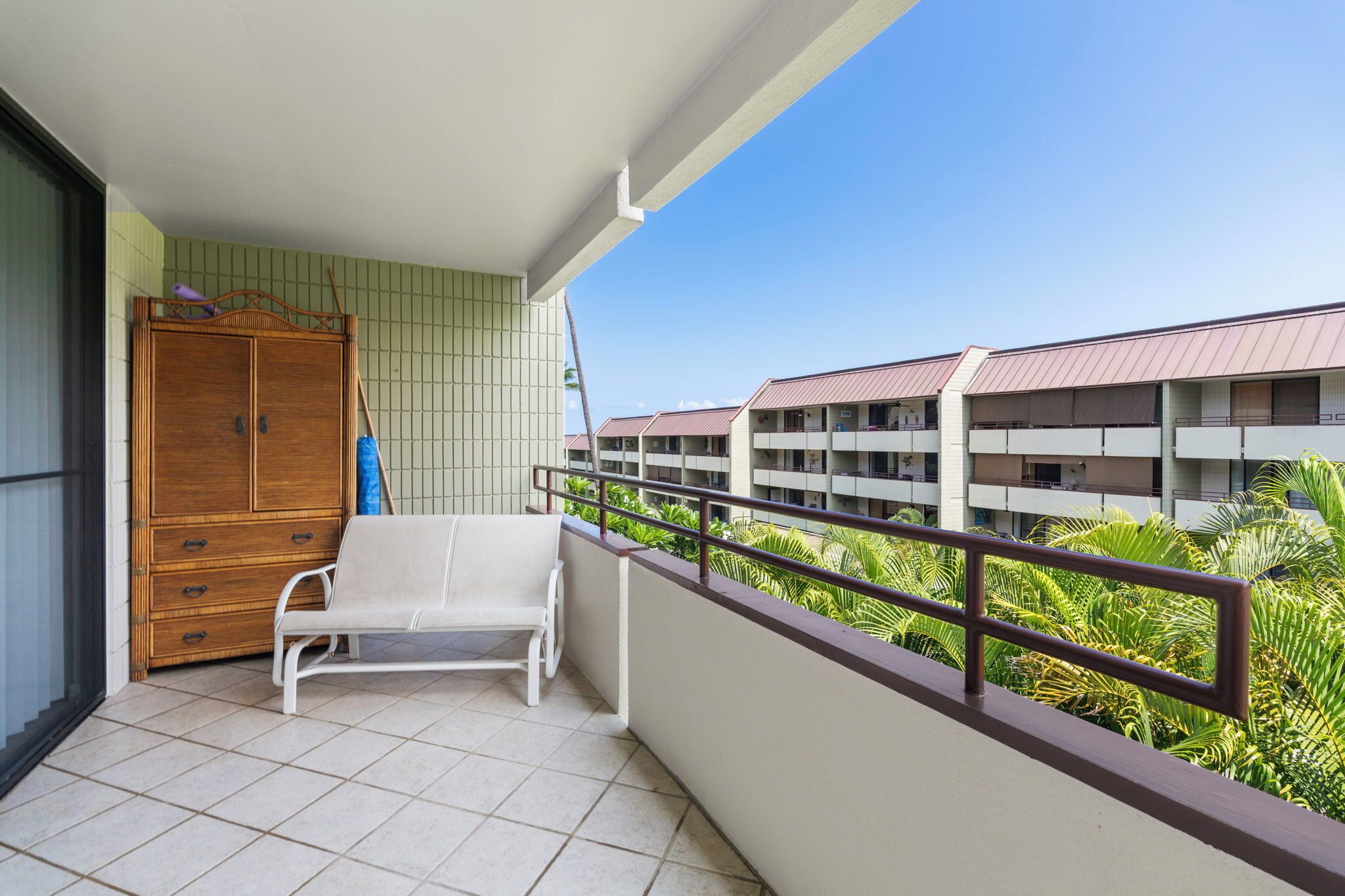 Un balcon avec une chaise blanche et une armoire en bois, donnant sur un immeuble doté de nombreux balcons et d’une végétation luxuriante.