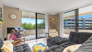 Una sala de estar moderna con un televisor de pantalla plana grande, sofás grises y un balcón con vistas a un edificio y zonas verdes.
